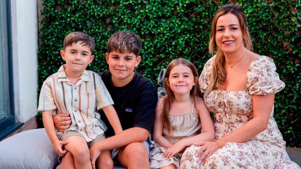 Three children sat next to their mum on a sofa. There is a hedge behind them. Jodanna wears a white and pink floral dress and has a young girl next to her. An older boy has Hudson on his lap.