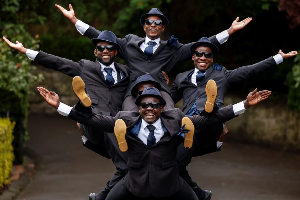 The Black Blues Brothers pose during a photocall for their Fringe show. Five men wearing matching dark suits, ties, hats and sunglasses are arranged in a stacked formation, with arms extended outward and legs raised.