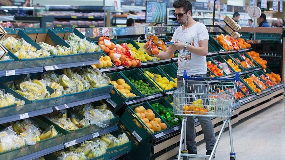 A man wearing a t-shirt uses a hand-held device to scan a net of oranges as he shops in the fruit and vegetable aisle of a Tesco supermarket