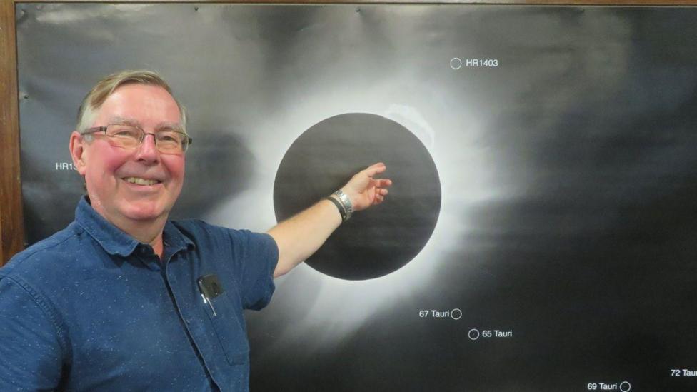 Jonathan Clough pointing at a diagram of a solar eclipse. He is the left of the image, wearing a blue shirt and smiling. 