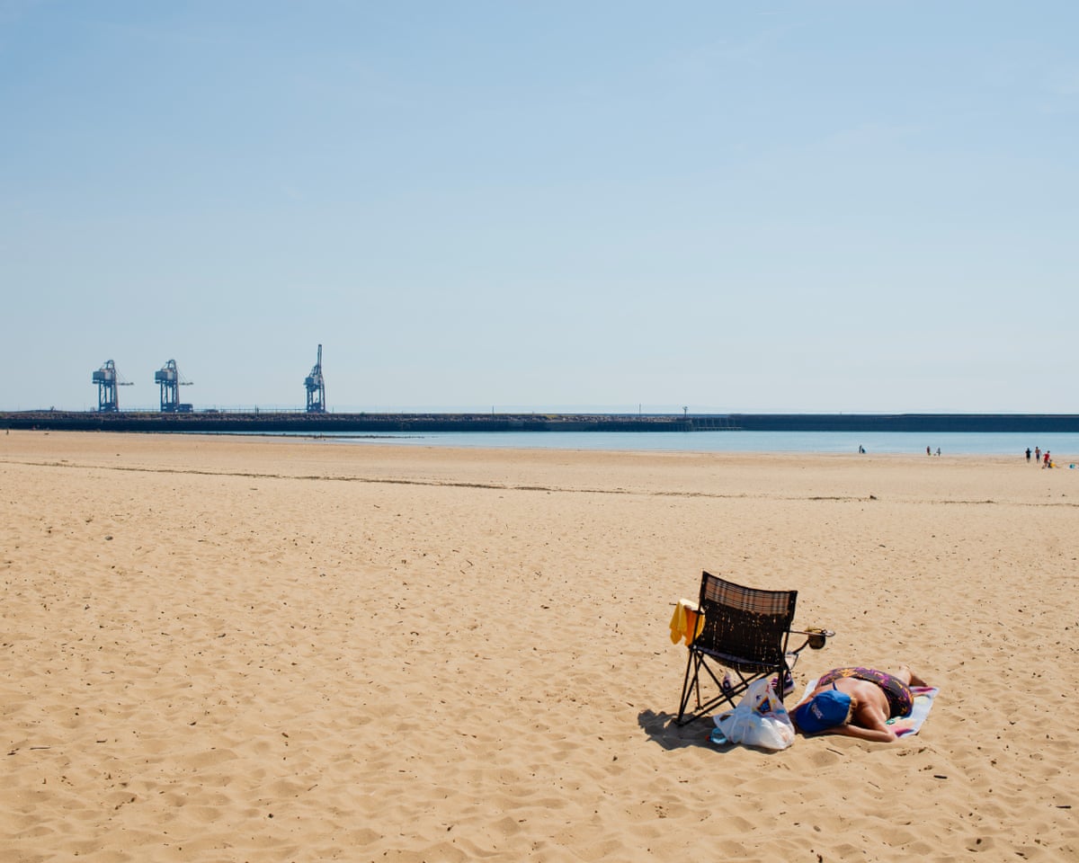 A woman sunbathing on a sandy beach with the works in the distance.