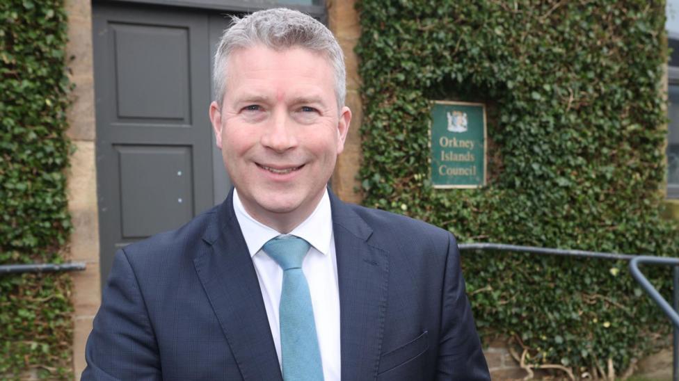 Oliver Reid, of Orkney Council, standing in front of council offices which are covered in ivy. He has short grey hair and is wearing a dark blue suit, white shirt and a light blue tie.