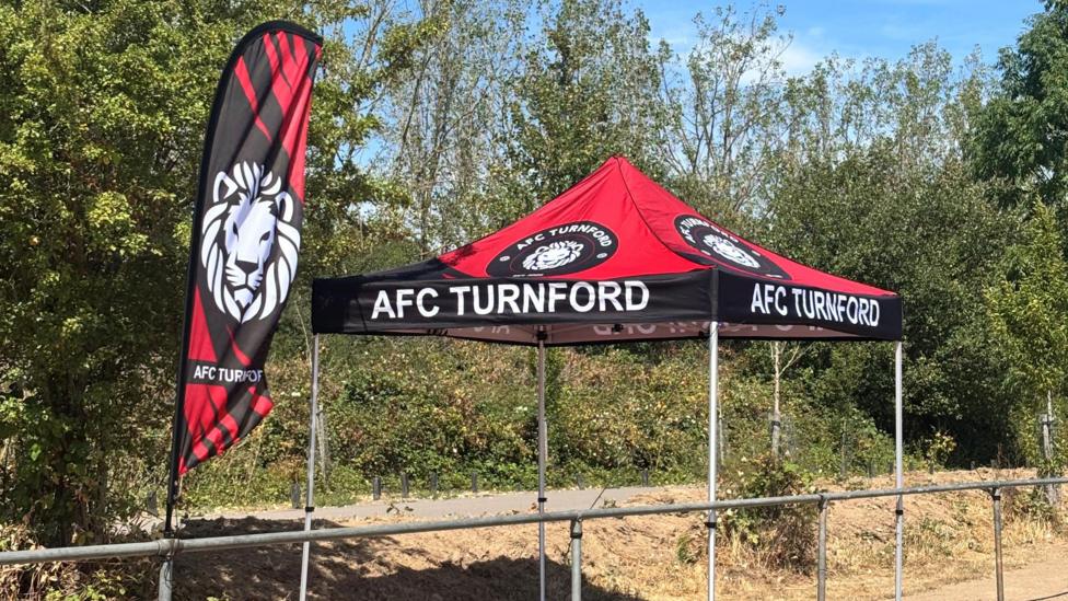 A flag and gazebo showing AFC Turnford, by a football pitch. It is made out of red and black colours. There are trees behind it and an area of grass, that is dried up.