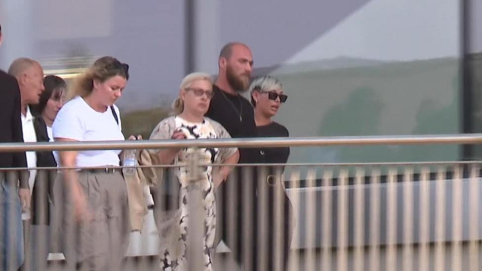 A group of people (men and women) walking past a building. The shot includes five people. 