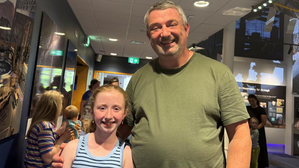 A girl and man pose for a photo, she has light hair and is wearing blue top, he has grey hair and is wearing green top