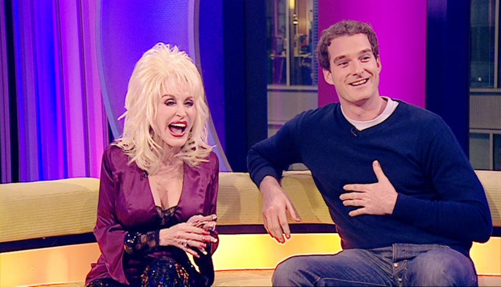 A screen shot of the one show with Dolly sitting on the yellow couch with Dan. she is wearing a purple low cut top and he is wearing a dark jumper and jeans. Both of laughing 