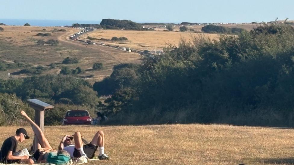 Several people sit and lie on dry grass on high ground near Beachy Head in East Sussex. A long queue of cars stretches along a road beyond them, with the sea visible in the distance.
