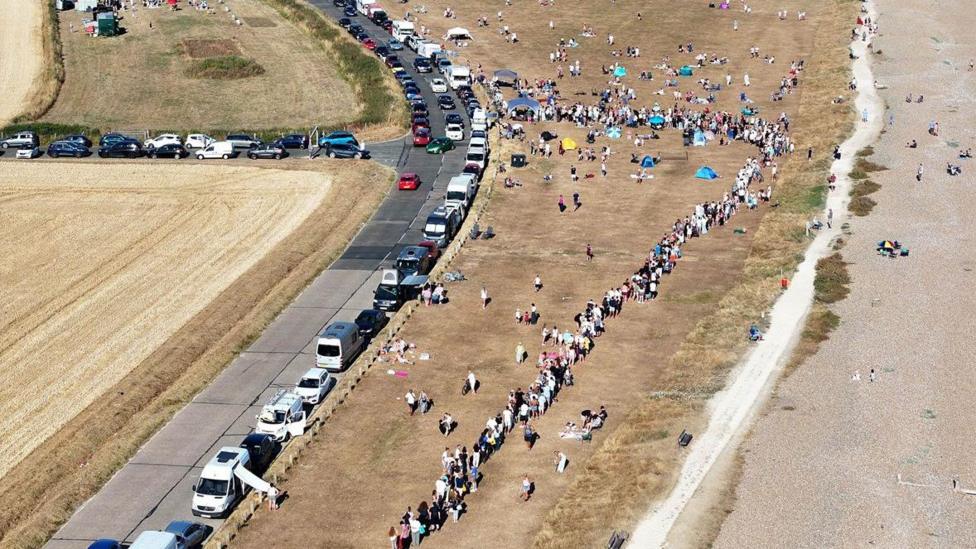 A large crowd gathers on a grassy coastal area near Goring-by-Sea in West Sussex. Long lines of parked cars stretch along a road beside the shoreline and a queue of people forms near viewing points.