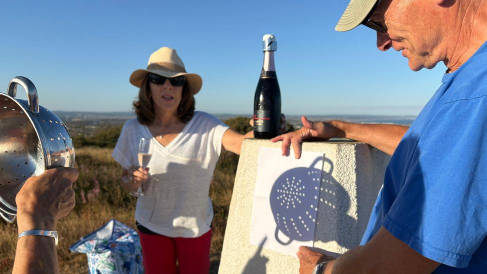 Glenda Whiles stands on the South Downs holding a drink while David Hearn holds a sheet of paper showing the shadow of a colander. Another person holds the colander in the foreground. Rolling countryside and distant views can be seen behind them.