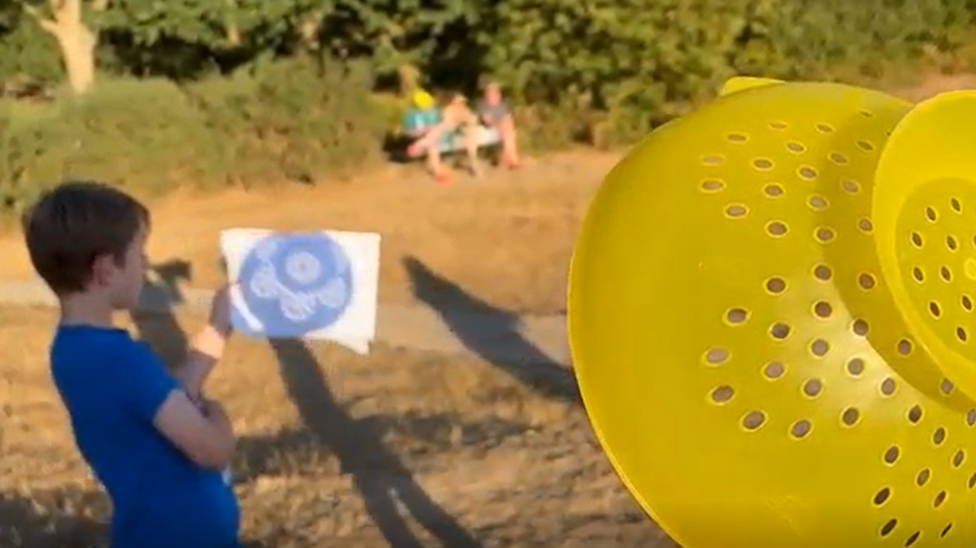 A bright yellow colander is held in the foreground while a child holds a piece of white card showing projected circles of sunlight. Other people are visible on dry grass in the background on Tunbridge Wells Common.