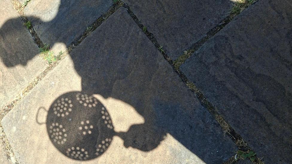 The shadow of a person holding a colander falls across paving stones in bright sunshine in Great Bookham, Surrey. The outline of the colander and its holes can be seen clearly on the ground.