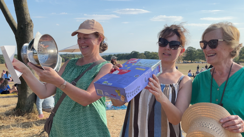 Three women: one with a colander and some paper and another with a cereal box.
