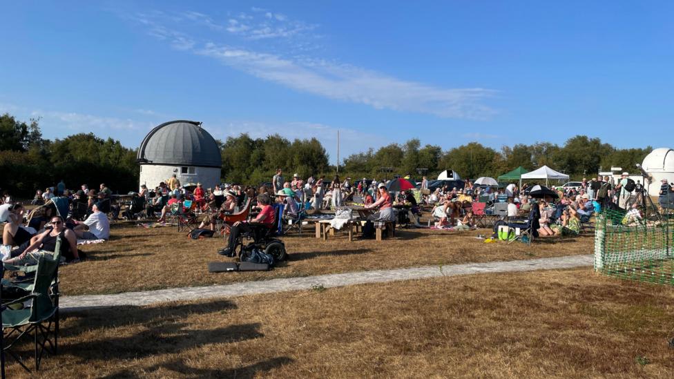 A picture of crowds gathering in a field. They are sat on a grass field.