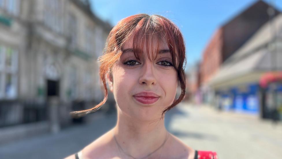 Charlotte stood on a quiet high street. She has a red fringe and looks at the camera.