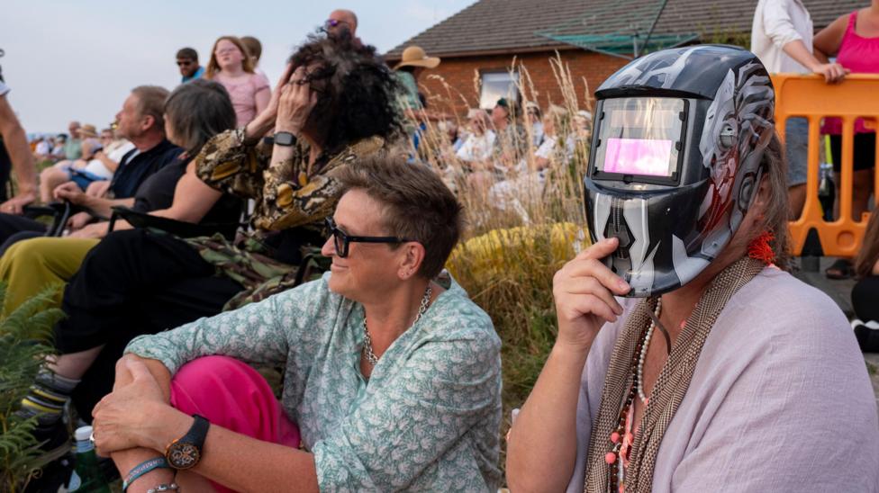 A woman holds a welder's mask to her face as people sit about in the sunshine looking skywards