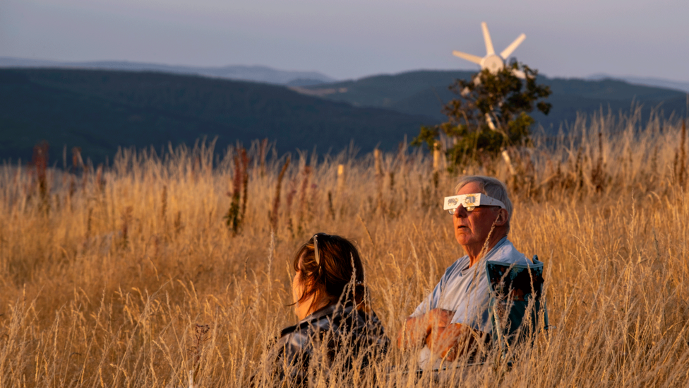 A man sits in camp chair, arms folded, looking skyward. He's wearing special glasses over his usual glasses. He is surrounded by long grass with Powys mountains in the background