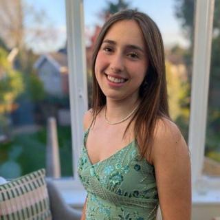 Rosa MacGowan smiles at the camera. She has long brown hair and has her hand in her jeans pocket. She is wearing a green vest top with sequins on it.