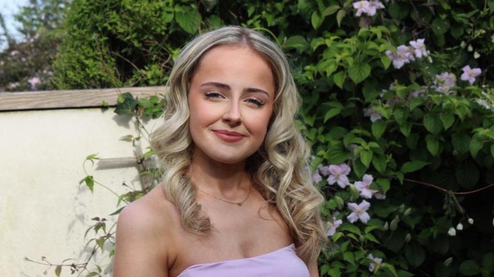 Caitlin, a young woman with wavy blonde shoulder-length hair wearing a pink dress in front of a bush with pink flowers on it.