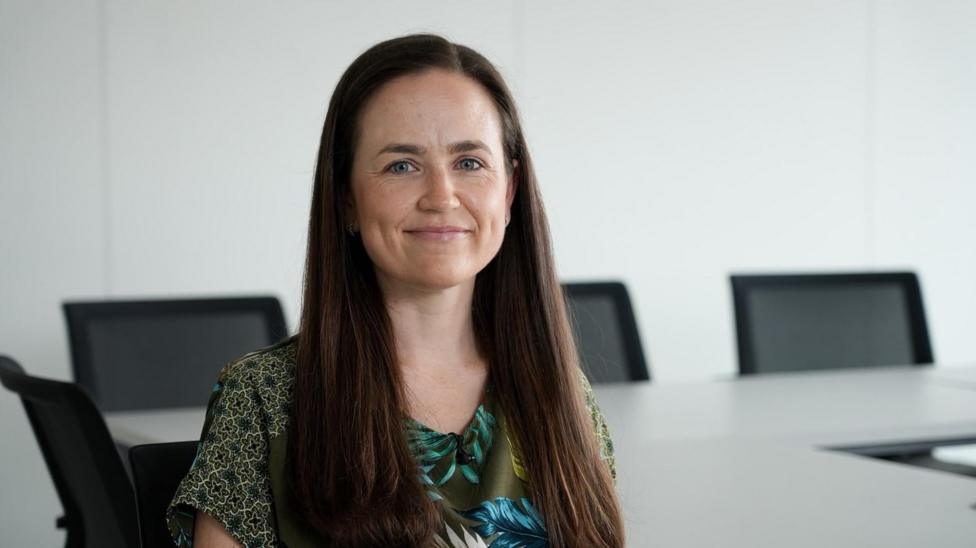 Bethan Evans with long dark hair is sitting in an office with white walls, a desk and black chairs in the background.