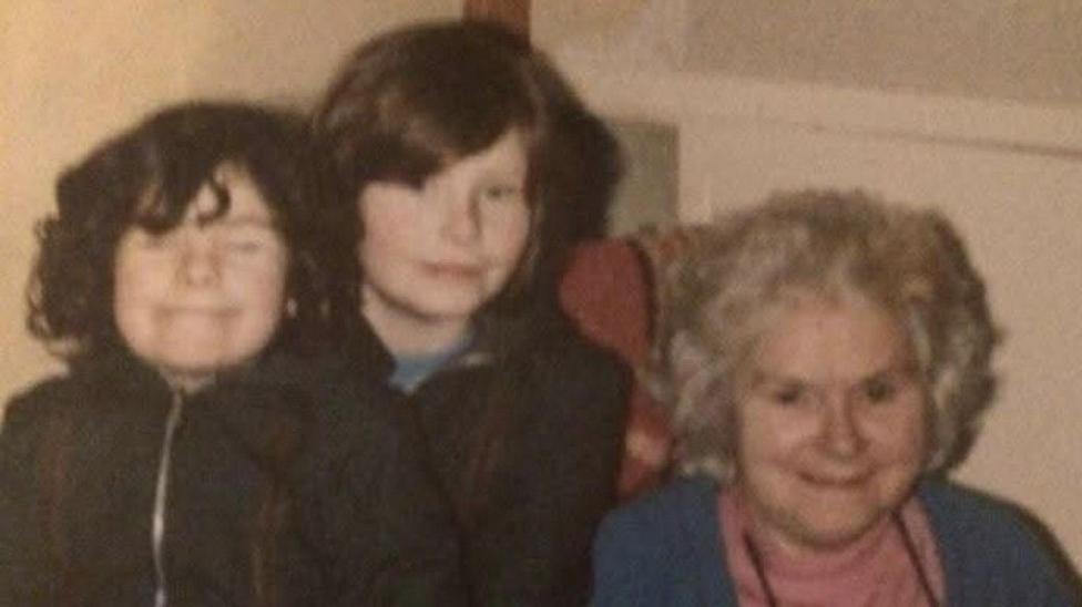 An old photograph of two young girls with dark hair and their grandmother. The girls are standing together next to an older woman who's sitting in a chair. They're all looking at the camera.