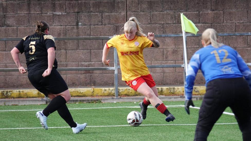A Stewarton Annick Ladies FC player on the ball, wearing the club kit of yellow short and red shorts. She is is taking on a defender on the other team, wearing an all black kit, and watched by the other team's goalkeeper, wearing a blue shirt and black jogging bottoms.