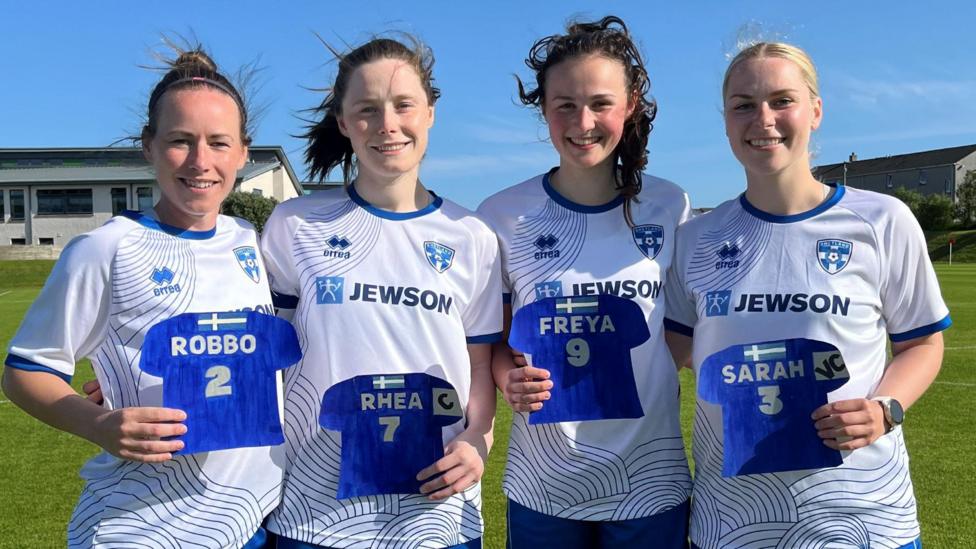 Four Shetland players standing together outside on a football pitch. They have their hair tied back and are wearing the white and blue colours of their club. They are also holding little replica football shirts with their names: Robbo, Rhea, Freya and Sarah.