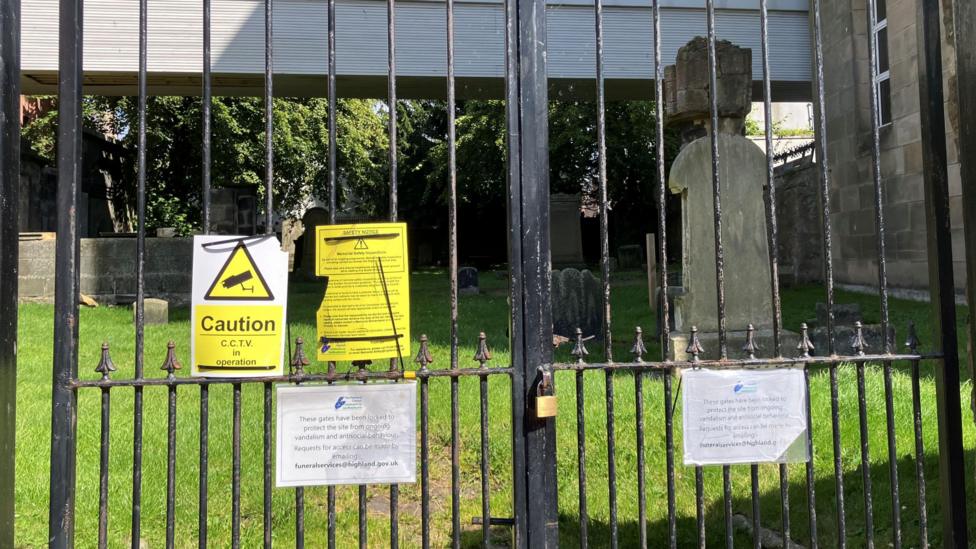 A gate made of black metal railings is closed and padlocked. There are signs explaining why the burial ground is closed. Behind the gate is an area of green grass bathed in sunlight and gravestones in the shadow of a nearby building.