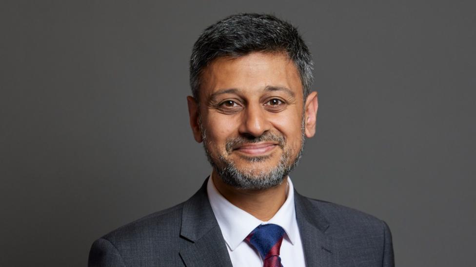 Dr Zubir Ahmed smiles, looking into the camera. He is wearing a white shirt, grey blazer and a blue and red tie