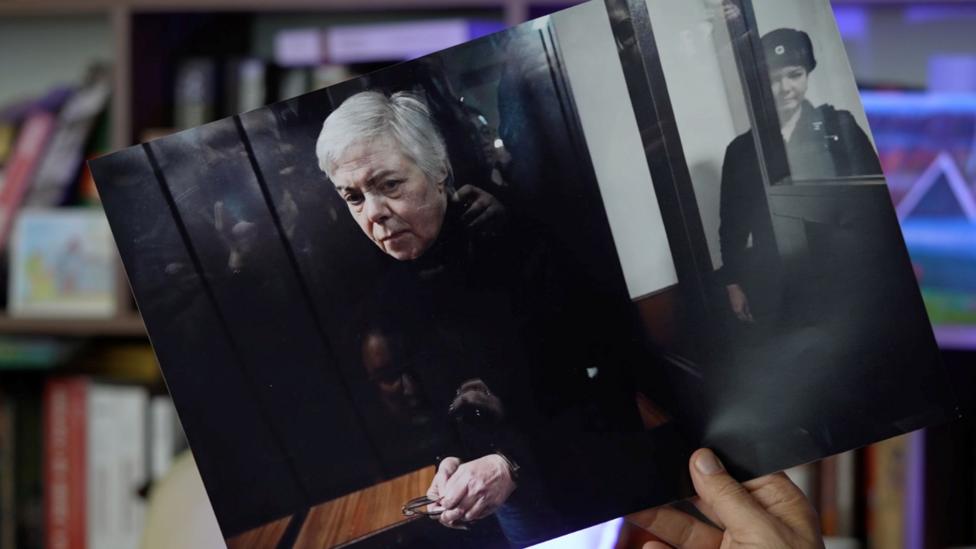 A hand holds a printed out photograph of Nadezhda Buyanova, a 68-year-old Ukrainian-born paediatrician in the dock at court, guarded.