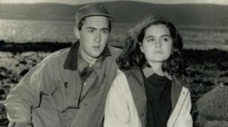 A black-and-white photograph shows Allan and Shirley seated among large rocks on a shoreline, with water and distant hills visible in the background. Both are dressed in outdoor clothing, creating a candid, natural scene in a rugged coastal setting.