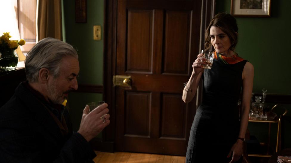 Two people hold drinks in a warmly lit room with dark green walls, polished wooden doors and traditional furnishings. One person sits in the foreground while the other stands nearby, creating a sense of distance and tension within the scene. Soft natural light from a window illuminates the interior, highlighting the rich wood tones and intimate atmosphere.