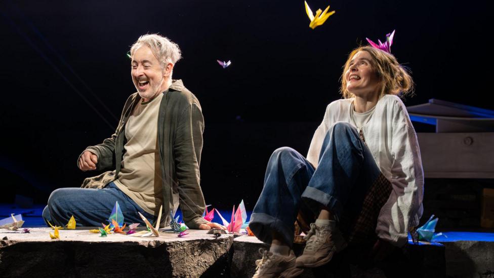 Two performers sit on the edge of a stage set, surrounded by brightly coloured origami cranes scattered around and suspended in the air. Dramatic spotlights illuminate the scene against a dark background, drawing attention to the vivid paper sculptures and casual costumes. The theatrical setting creates a striking contrast between the simple staging and the whimsical, dreamlike display of floating paper birds.