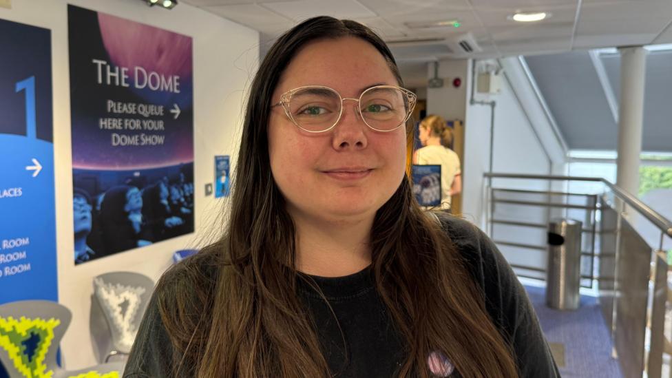 Cathleen is a young woman with long, dark hair and silver glasses. She is smiling at the camera. She is wearing a black t-shirt and standing in front of a sign that points towards the planetarium's dome show room.