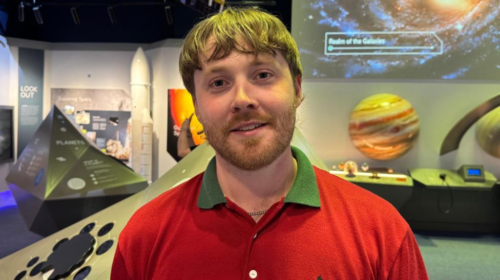 A head and shoulders picture of Kev. He has short, red hair and a beard. He is wearing a red polo t-shirt with a green collar. He is smiling at the camera. Behind him are different displays in the Planetarium, including some model displays of planets and stars.