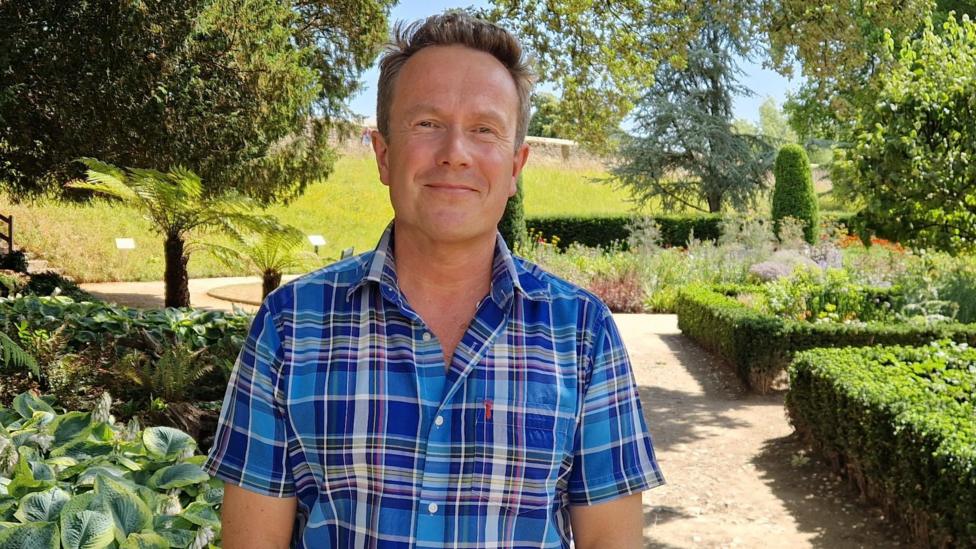 Wearing a short sleeved blue, checked shirt, James smiles at the camera. He stands on a path within a well kept garden, with a small hedge lining the path on his left and other plants on his left. Several trees also surround him.