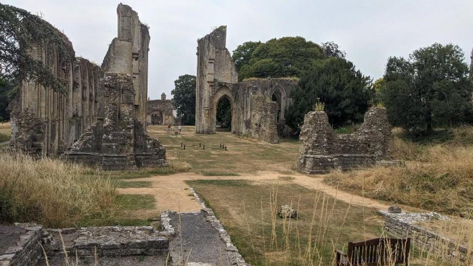 The photo shows the ruins of Glastonbury Abbey. A grassy lawn fills the middle of the image, with stone arches to its left and right. The grass is largely brown.