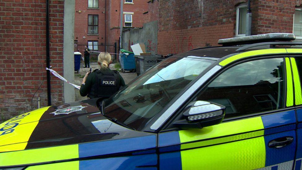 A blonde police officer stands near a police car
