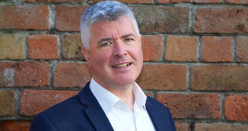 A man with grey hair, wearing a blue suit jacket and open-necked shirt, stands in front of a brick wall