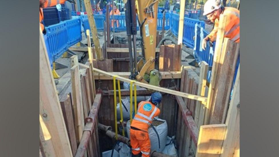 Engineers carrying out repair work in a deep excavated trench around a large water main. A worker in high-visibility orange protective clothing stands at the bottom of the shored excavation beside exposed pipework and repair materials, while another worker operates machinery from the surface.