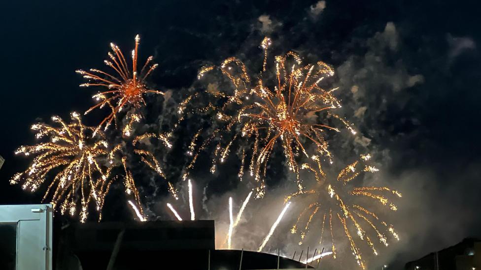 Fireworks bursting in the night sky above buildings near a waterfront.