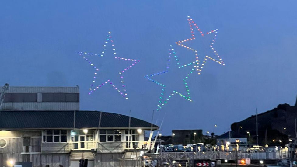 Colourful star-shaped drone lights displayed in the sky above a harbour with boats and waterfront buildings.