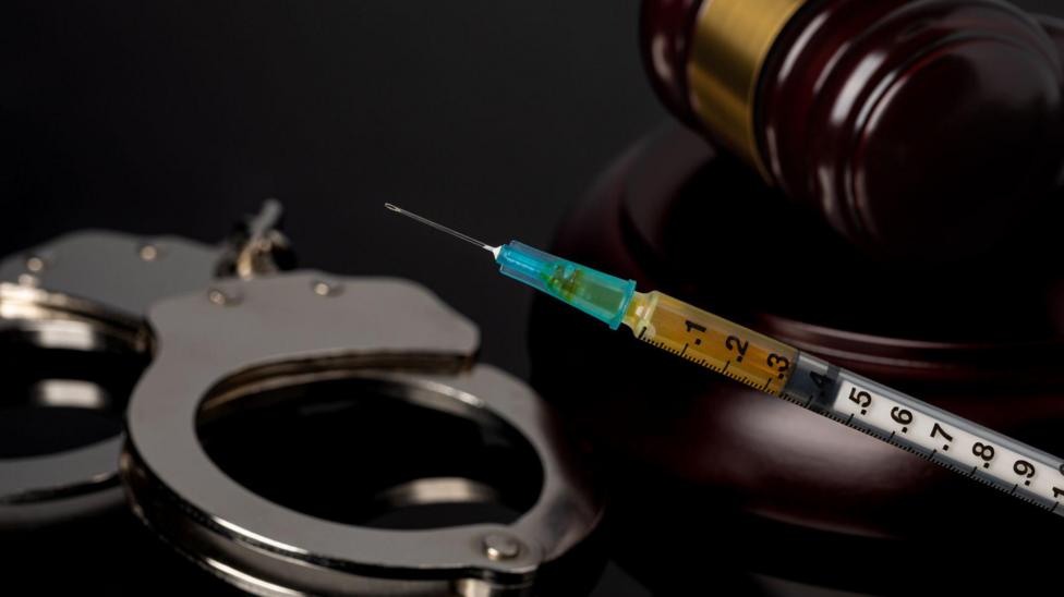 A stock image that shows handcuffs, a gavel and a syringe