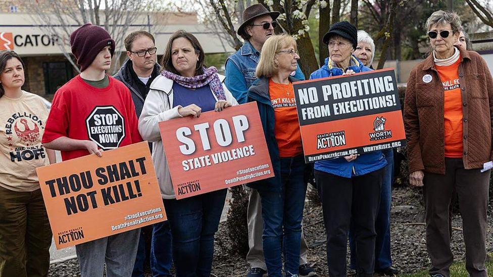 A group of protesters in coats and hats hold signs that say