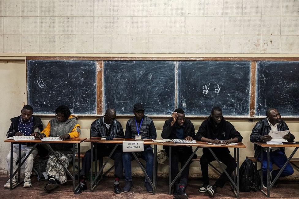 A group of officials sit behind trestle tables. Behind them are four school blackboards fixed to the wall.