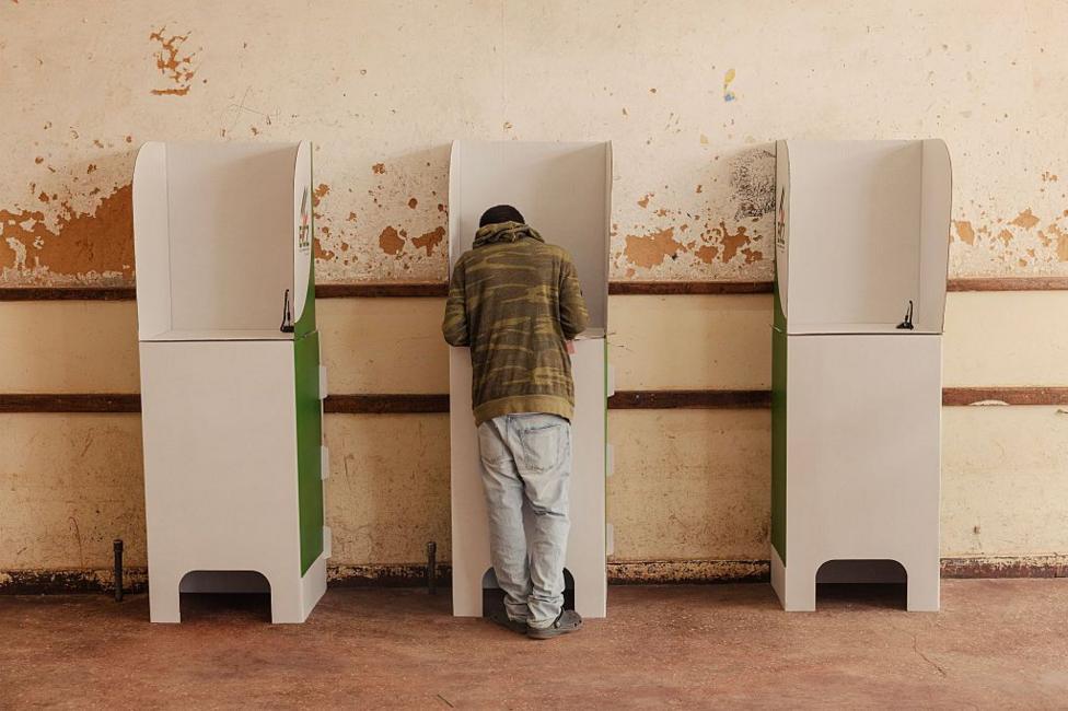 A voter at a cardboard ballot booth has their back to the camera. The voter is in the middle of three ballot booths set against a scuffed wall.