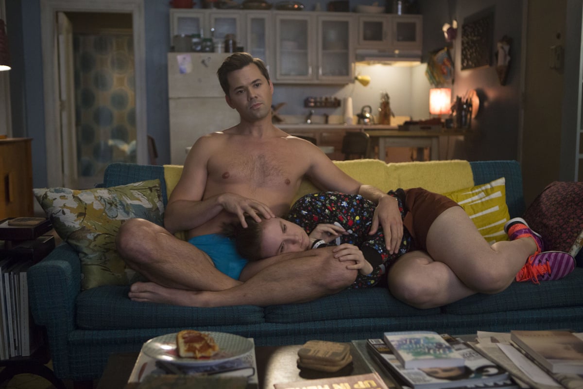 A topless man consoles a woman with her head in his lap while sitting on a sofa