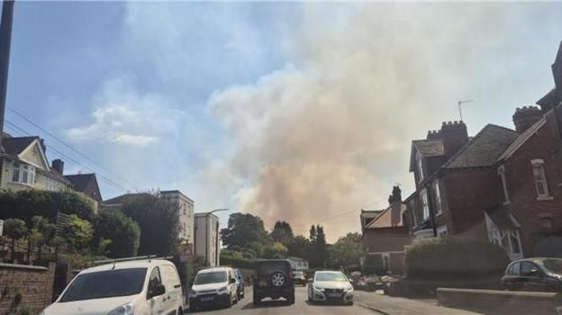 A large plume of smoke can be seen in the distance, billowing above a residential street in the foreground with cars parked either side.