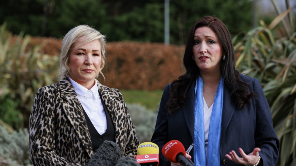 Michelle O'Neill, a woman with blonde hair, in a leopardskin jacket, with white blouse and black top, alongside Emma Little-Pengelly, a woman with dark hair, a white blouse, light blue scarf and a dark blue jacket.