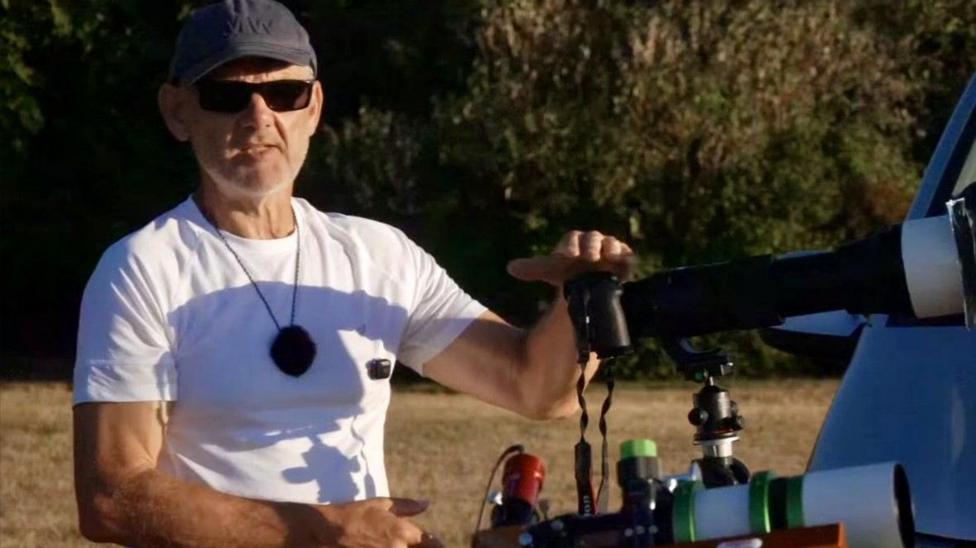 A man in a white T-shirt, and wearing a grey baseball cap and sunglasses, rests his left hand on a telescope. He is standing on a grassy area with hedging behind him.