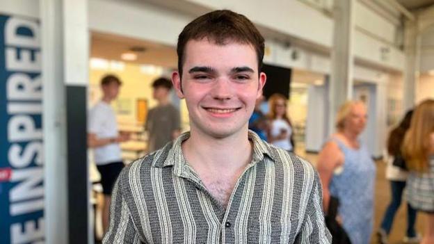 Stephen has short hair and wears a grey and white striped shirt. He is smiling and holding papers with his results on. In the background people are milling about in a school hall